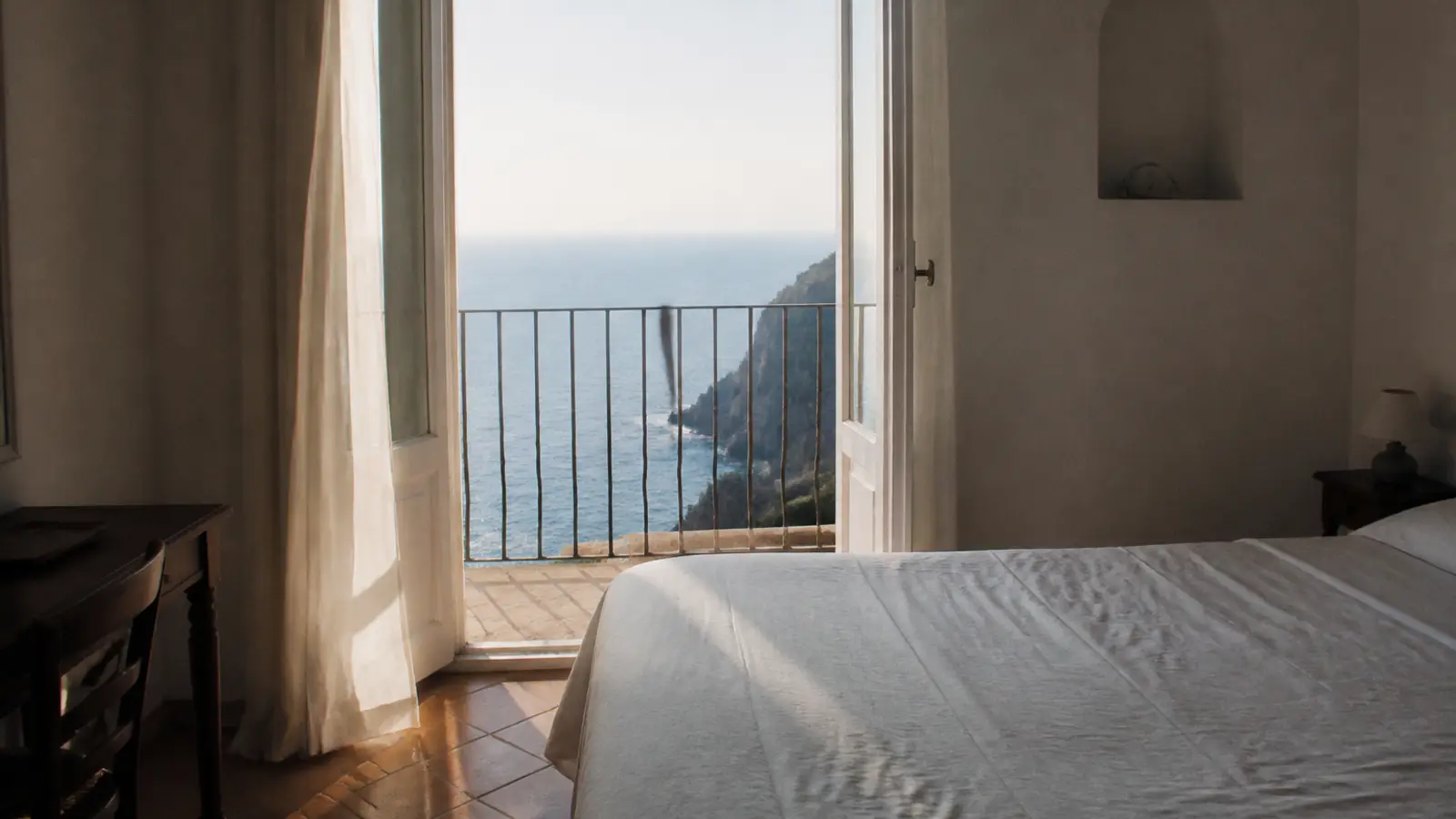 Room with a white bed and doors open onto a balcony over the sea